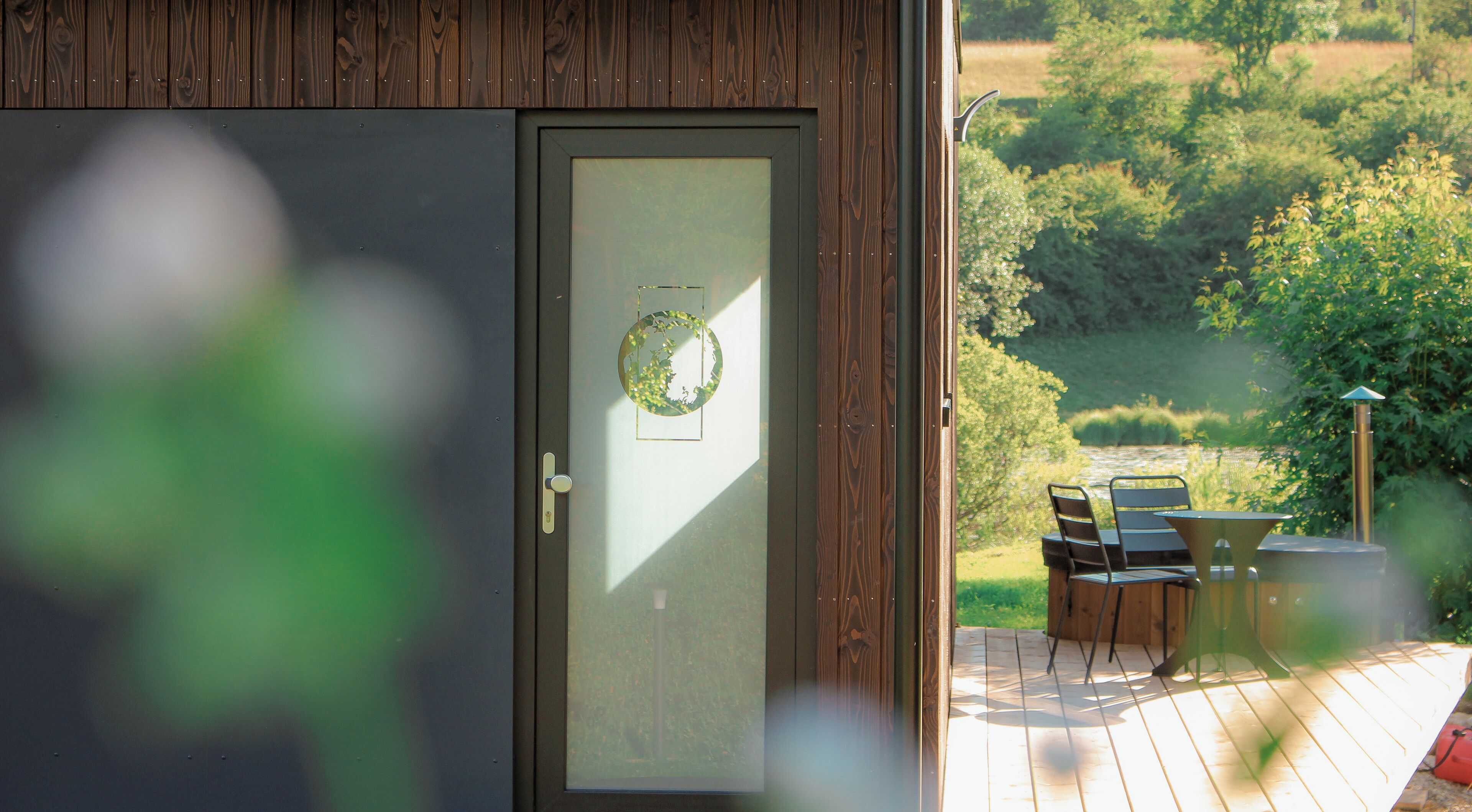 A wooden door with a frosted glass panel and a circular decoration. Outside, a deck with chairs and a table is visible, overlooking a green landscape.