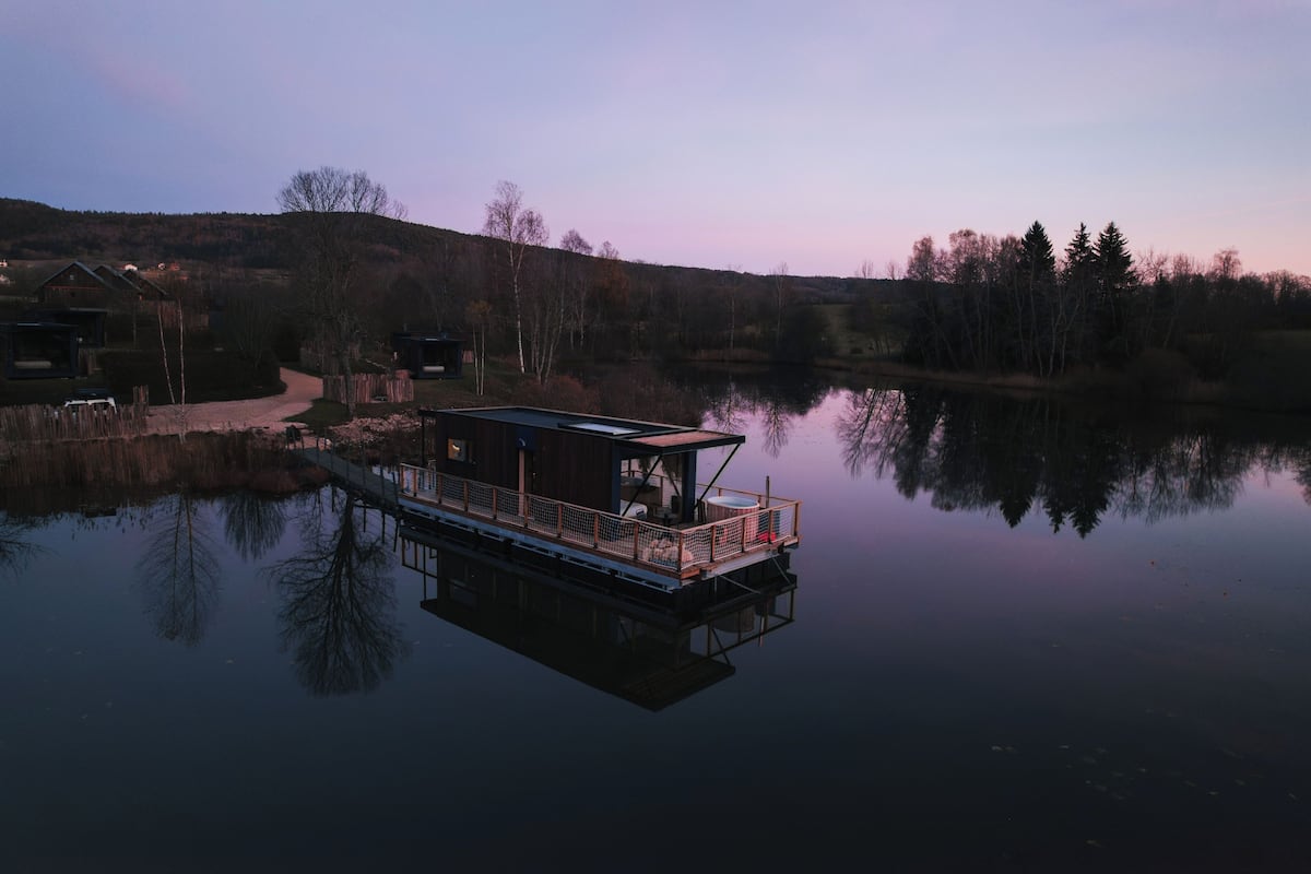 A floating house with a deck and a small cabin, surrounded by a calm lake and trees.