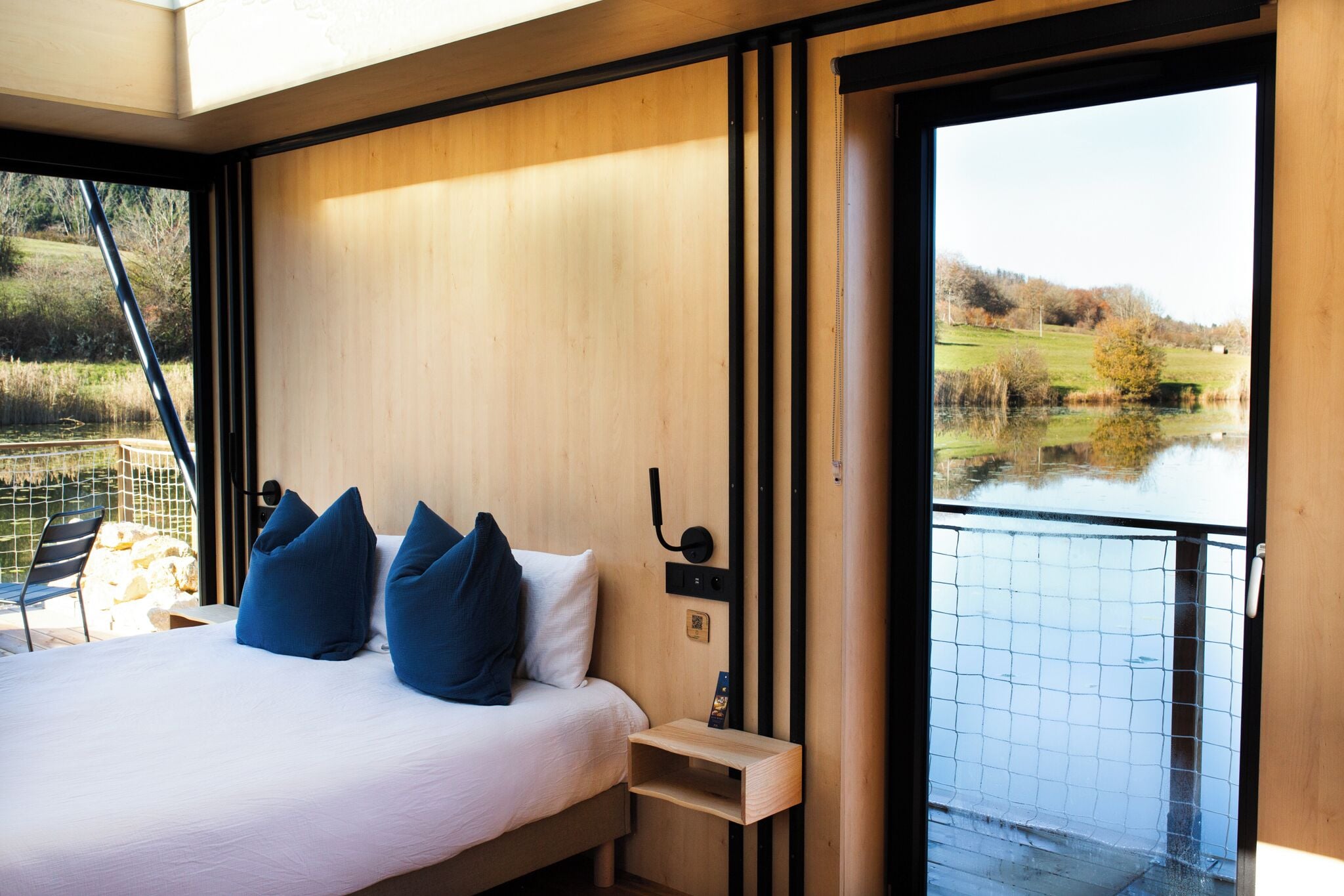 Quarto com cama, almofadas, mesa de cabeceira e vista para um lago.