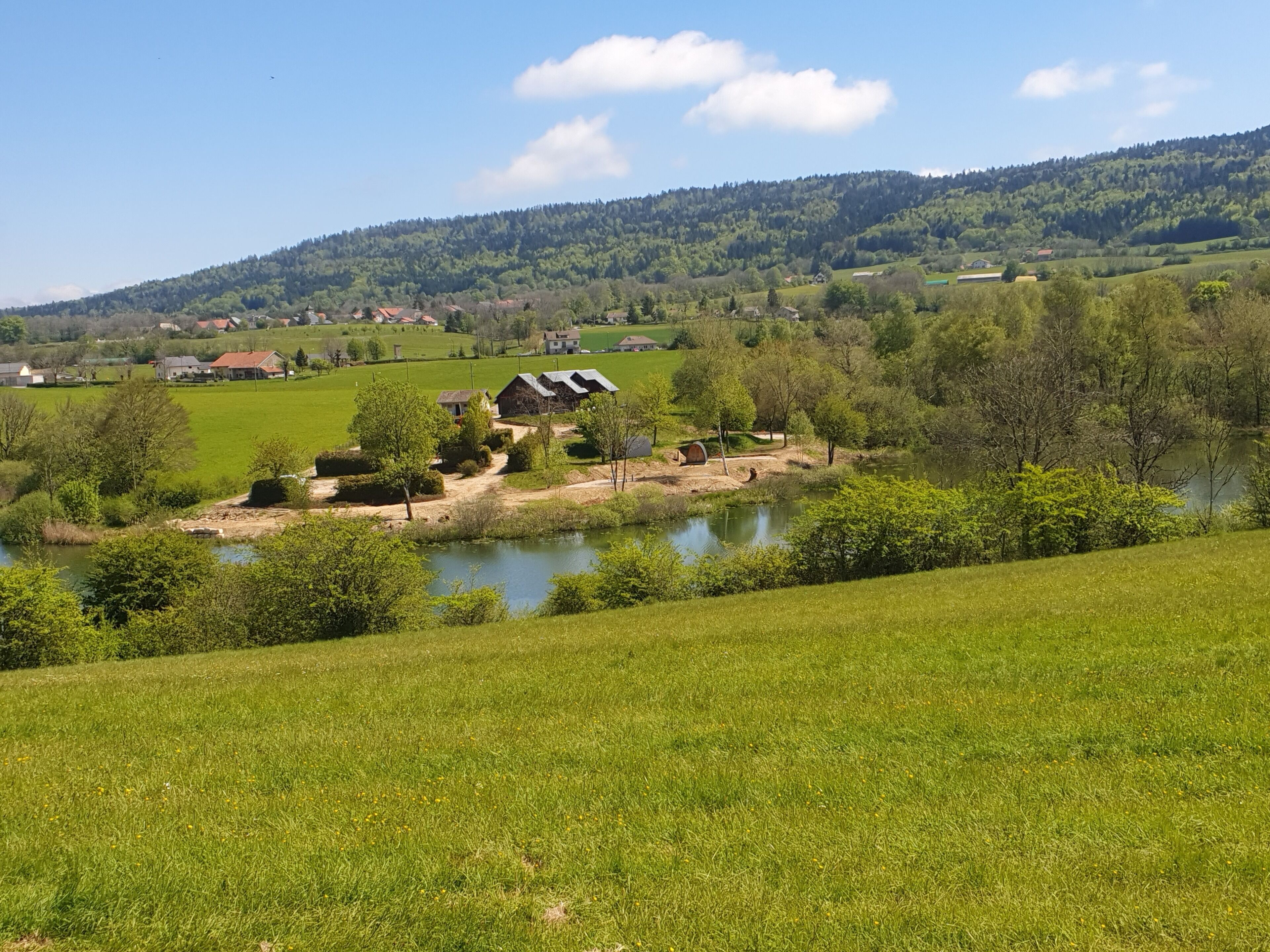 A rural landscape with a small house near a pond, surrounded by greenery and hills.
