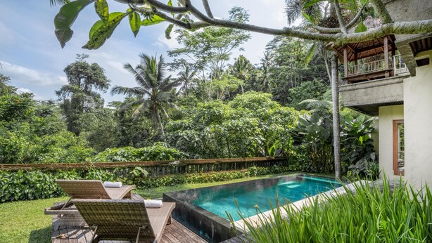 Un área de piscina con sillones reclinables y una terraza de madera, rodeada de exuberante vegetación y un edificio de dos pisos.