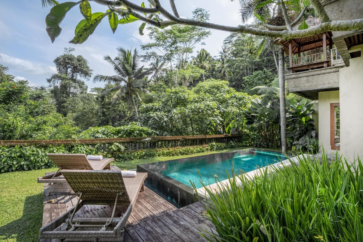 Un área de piscina con sillones reclinables y una terraza de madera, rodeada de exuberante vegetación y un edificio de dos pisos.