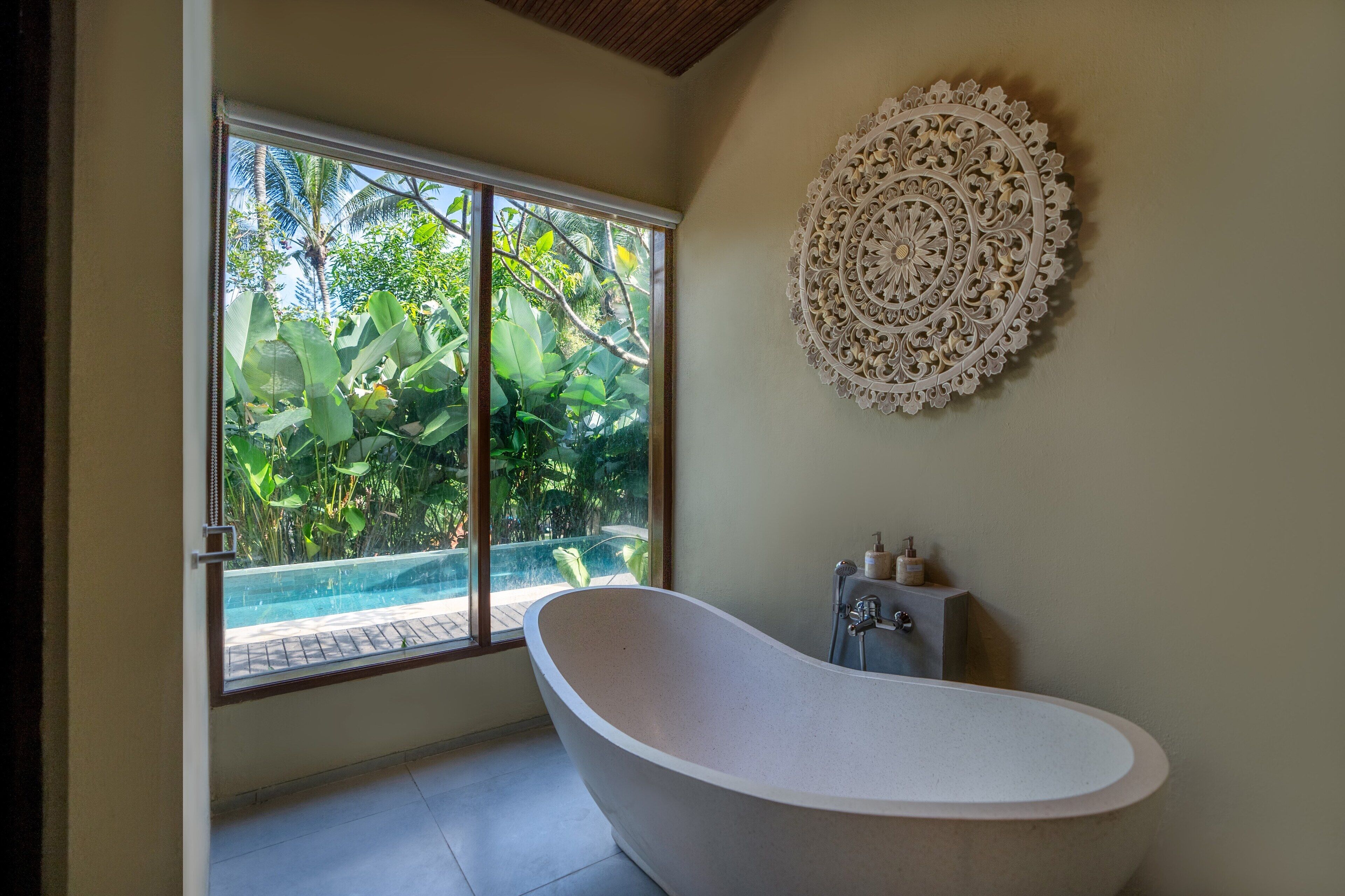 A modern bathroom with a freestanding bathtub, a large window with a view of lush greenery, and a decorative wall piece.