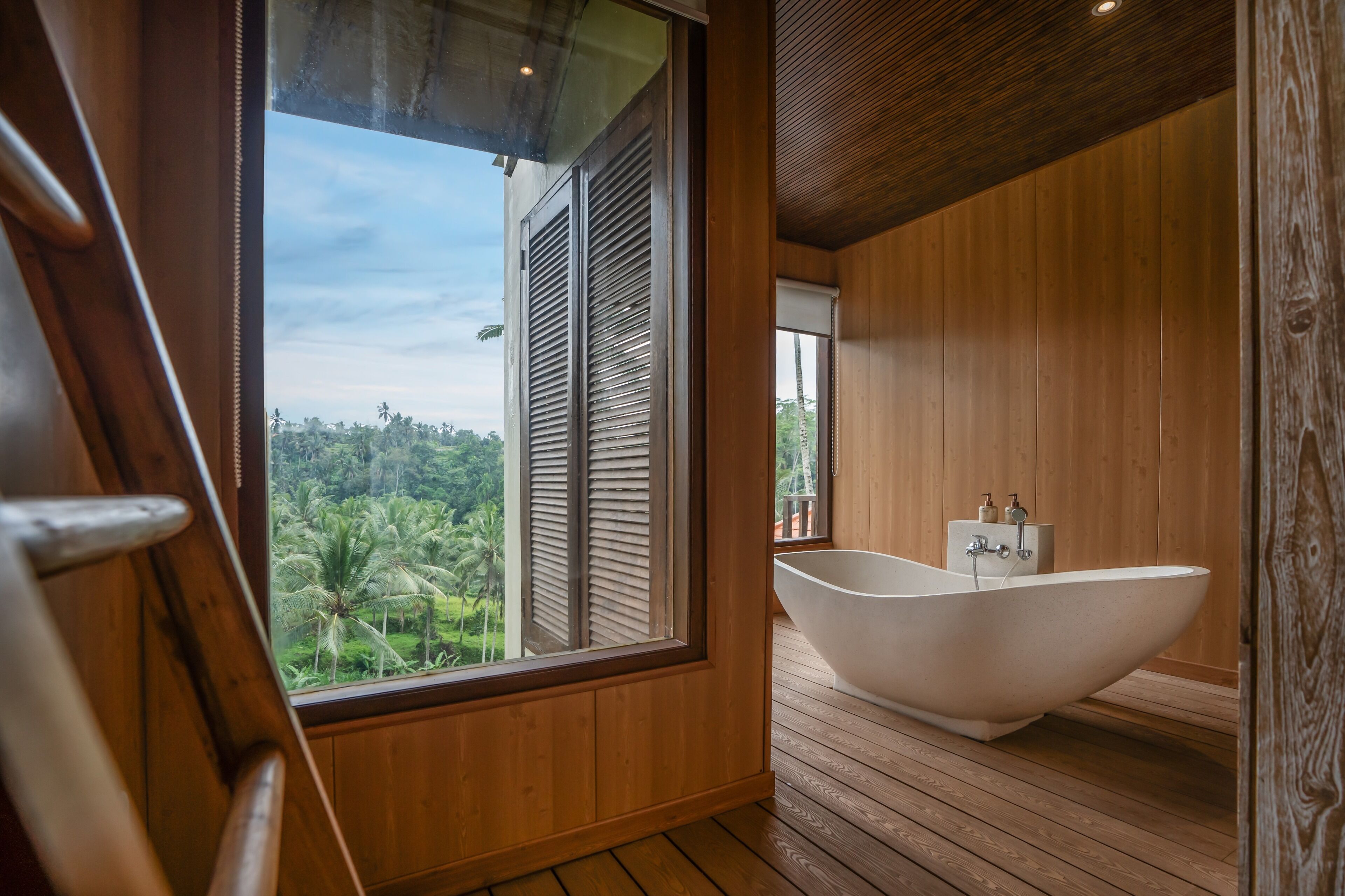 A wooden-floored bathroom with a freestanding bathtub and a large window offering a view of palm trees.
