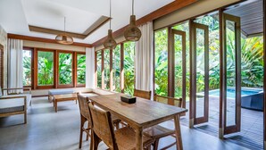 A spacious room with a large wooden dining table, wicker chairs, and a view of a lush garden and pool through large sliding glass doors. - Tjendana Kanaka Jungle Resort Ubud (Payangan)