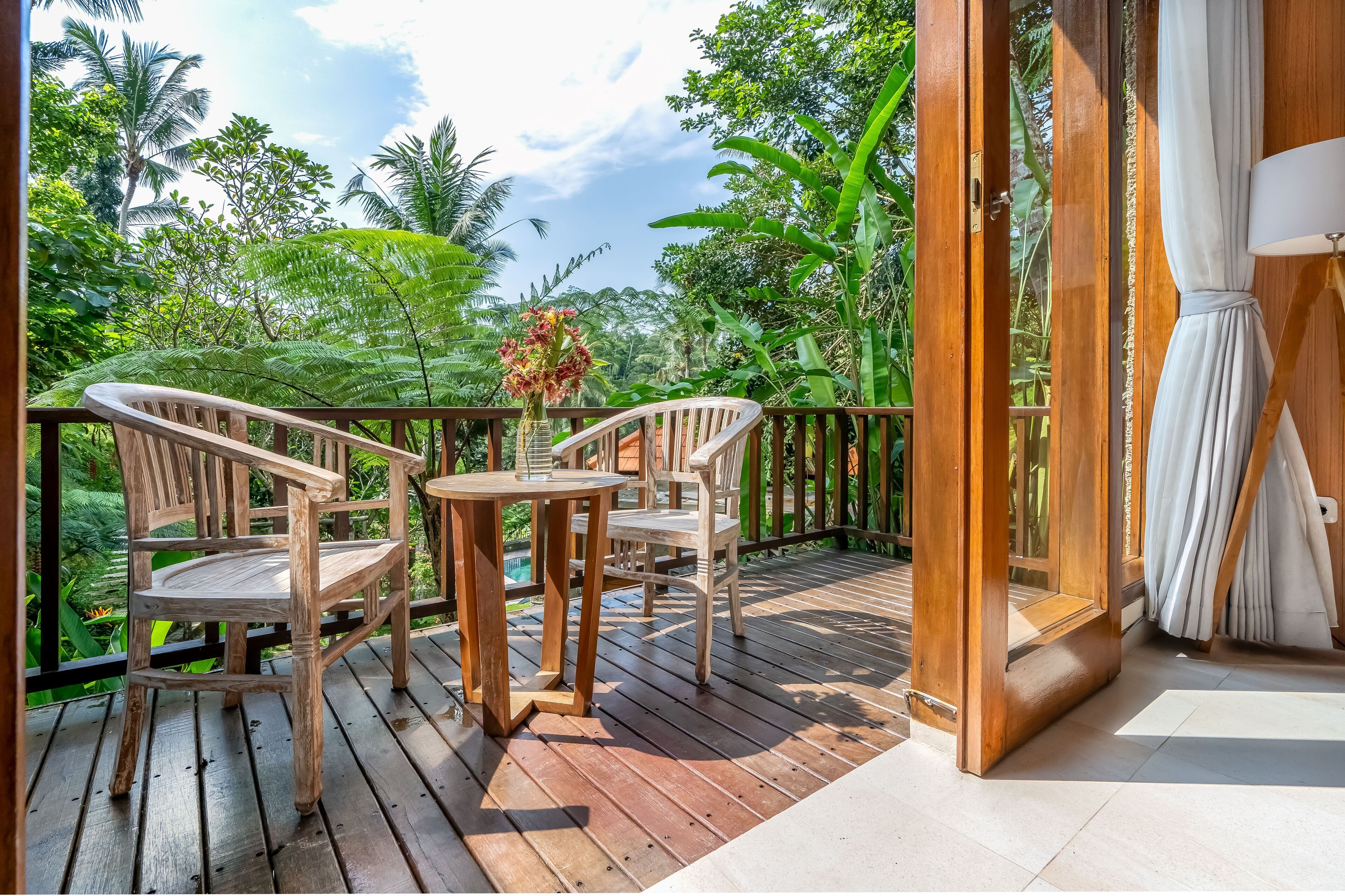 A wooden deck with a table and chairs, surrounded by lush greenery and a view of the sky.