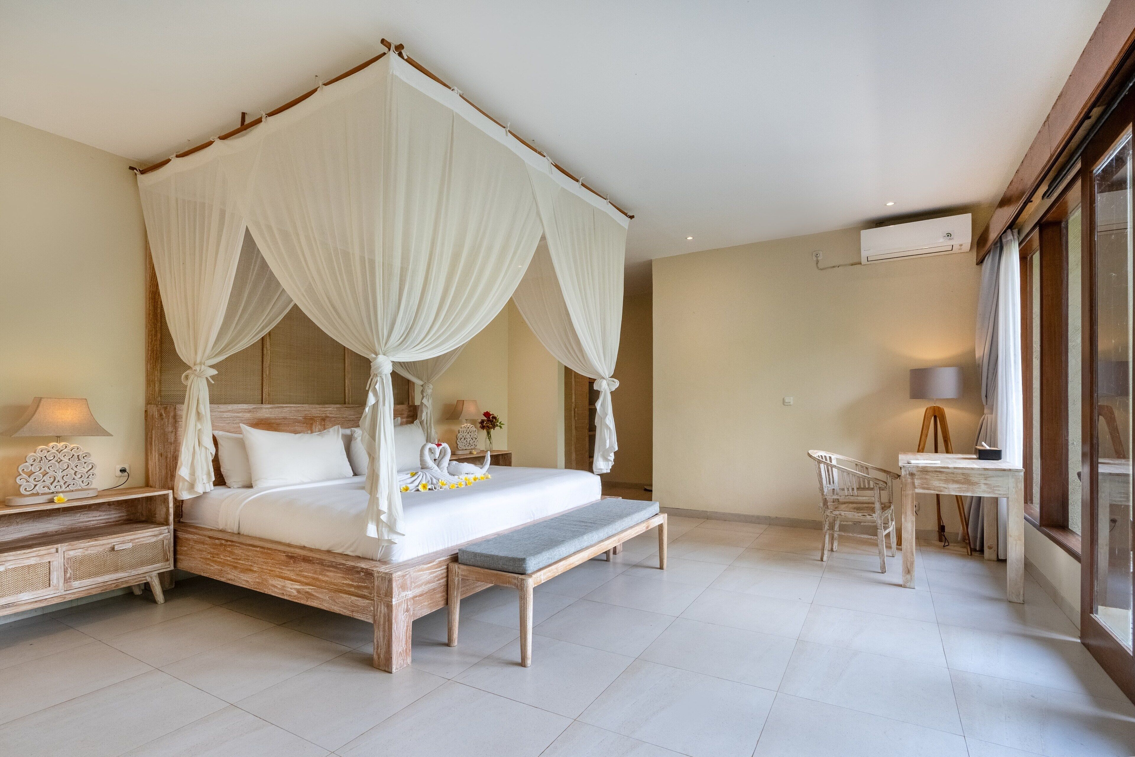 A four-poster bed with a canopy, a bedside table with a lamp, and a chair in a well-lit room with large windows.