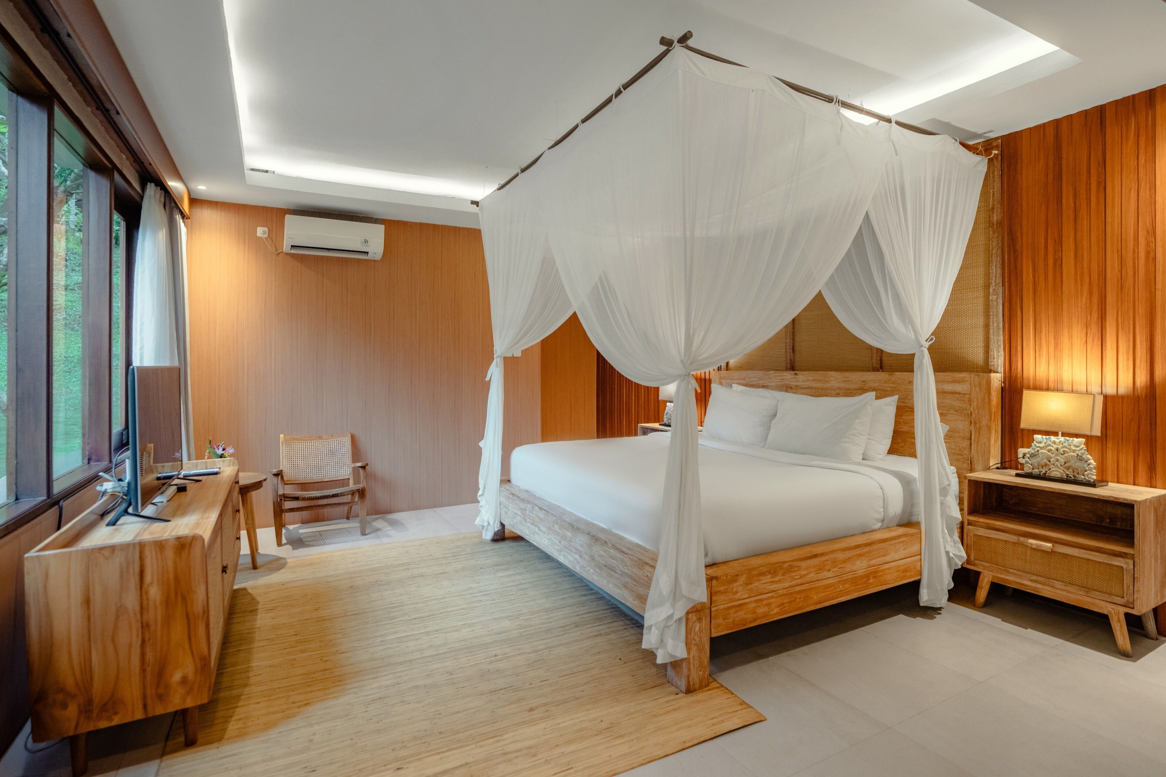 A bedroom with a four-poster bed, wooden furniture, a desk with a chair, and a nightstand with a lamp.