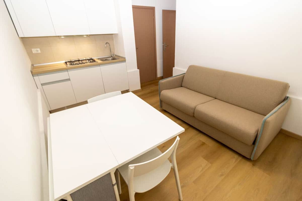 Una kitchenette compacta con una encimera blanca, estufa y pileta. Se observa un sofá color beige y una mesa de comedor blanca con una silla.