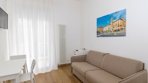 A modern living room with a beige sofa, a white table, and a white chair. There is a radiator and a painting of a building on the wall. - DimmiDiRimini (Rimini)