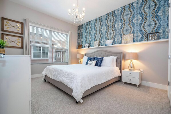 A bedroom with a large bed, a nightstand, a chandelier, and a patterned wallpaper. - Cozy Villa 5 min to Disney (Kissimmee)