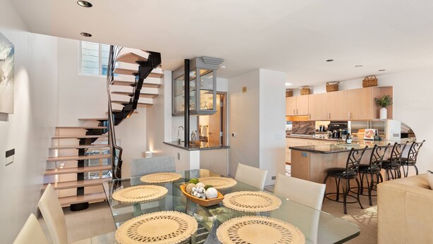 Una cocina moderna con una isla central, un comedor con una mesa de vidrio y individuales tejidos, y una escalera en espiral que conduce a un nivel superior.