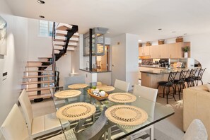 Una cocina moderna con una isla central, un comedor con una mesa de vidrio y individuales tejidos, y una escalera en espiral que conduce a un nivel superior.