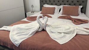 Una cama bien hecha con una toalla dispuesta en forma de corazón, un cabecero capitonado gris y almohadas marrones.
