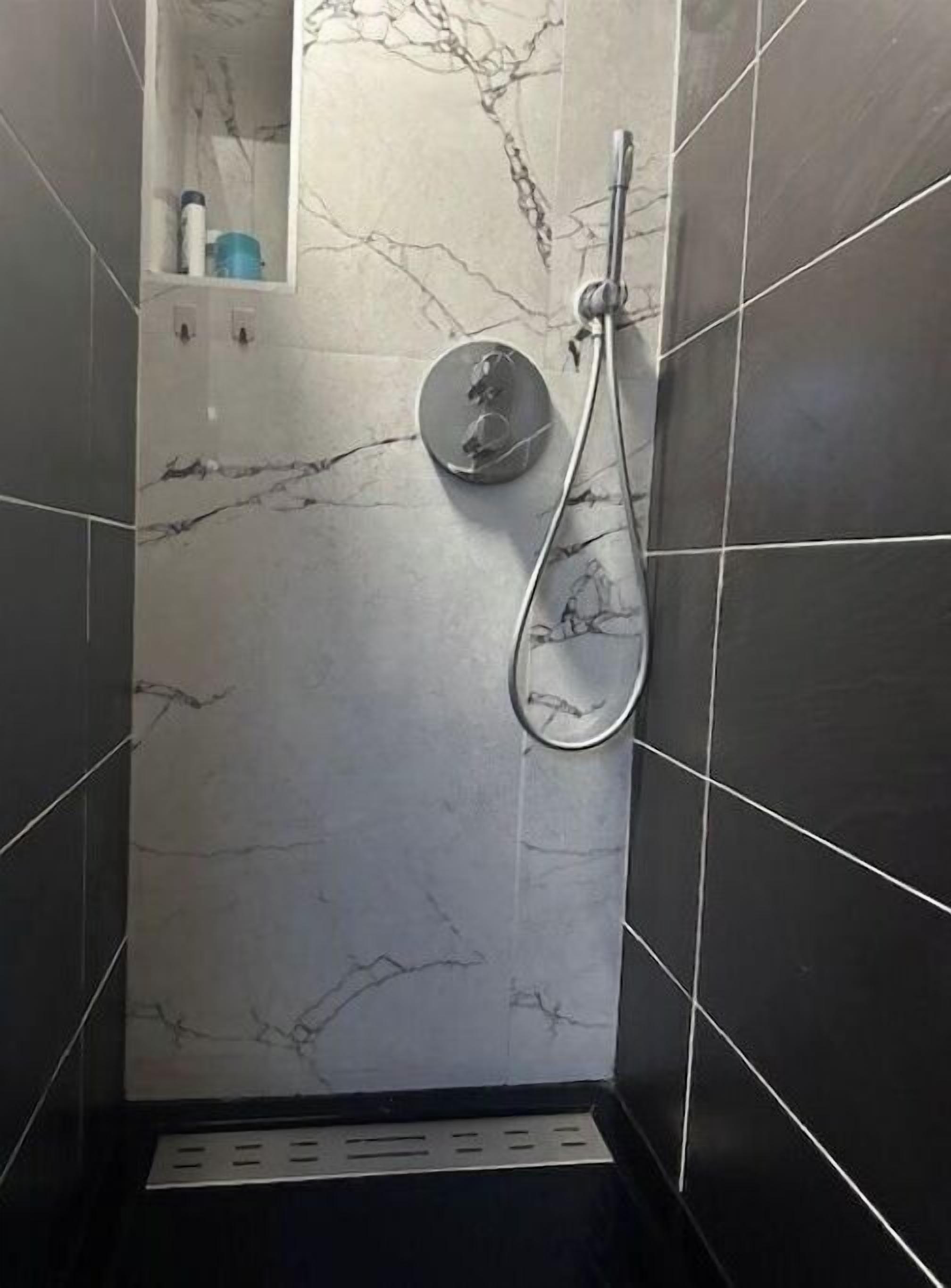 Une douche avec un mur en marbre, un pommeau de douche à main et une petite étagère pour les articles de toilette.