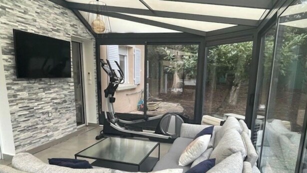 Een moderne woonkamer met een glazen uitbouw, grijze banken, een loopband en een flatscreen televisie.