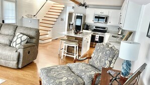 A spacious living room with a grey sofa, patterned armchair, and a wooden coffee table. A staircase leads to a kitchen with white cabinets and modern appliances. - T’is Our Cottage:Modern Lakefront fall colors & football relax Just off toll Rd  (Howe)