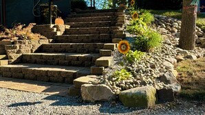 A house with a stone staircase leading to a porch, surrounded by a garden with sunflowers and a flag. - T’is Our Cottage:Modern Lakefront fall colors & football relax Just off toll Rd  (Howe)