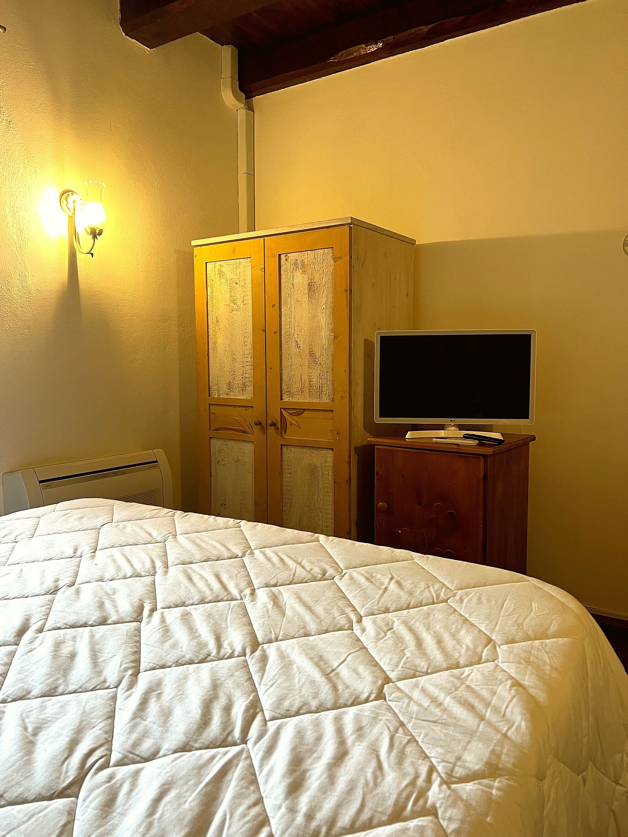 Ein Zimmer mit einem Bett, einem Holzschrank, einem Fernseher auf einer Holzkommode und einer Wandleuchte.