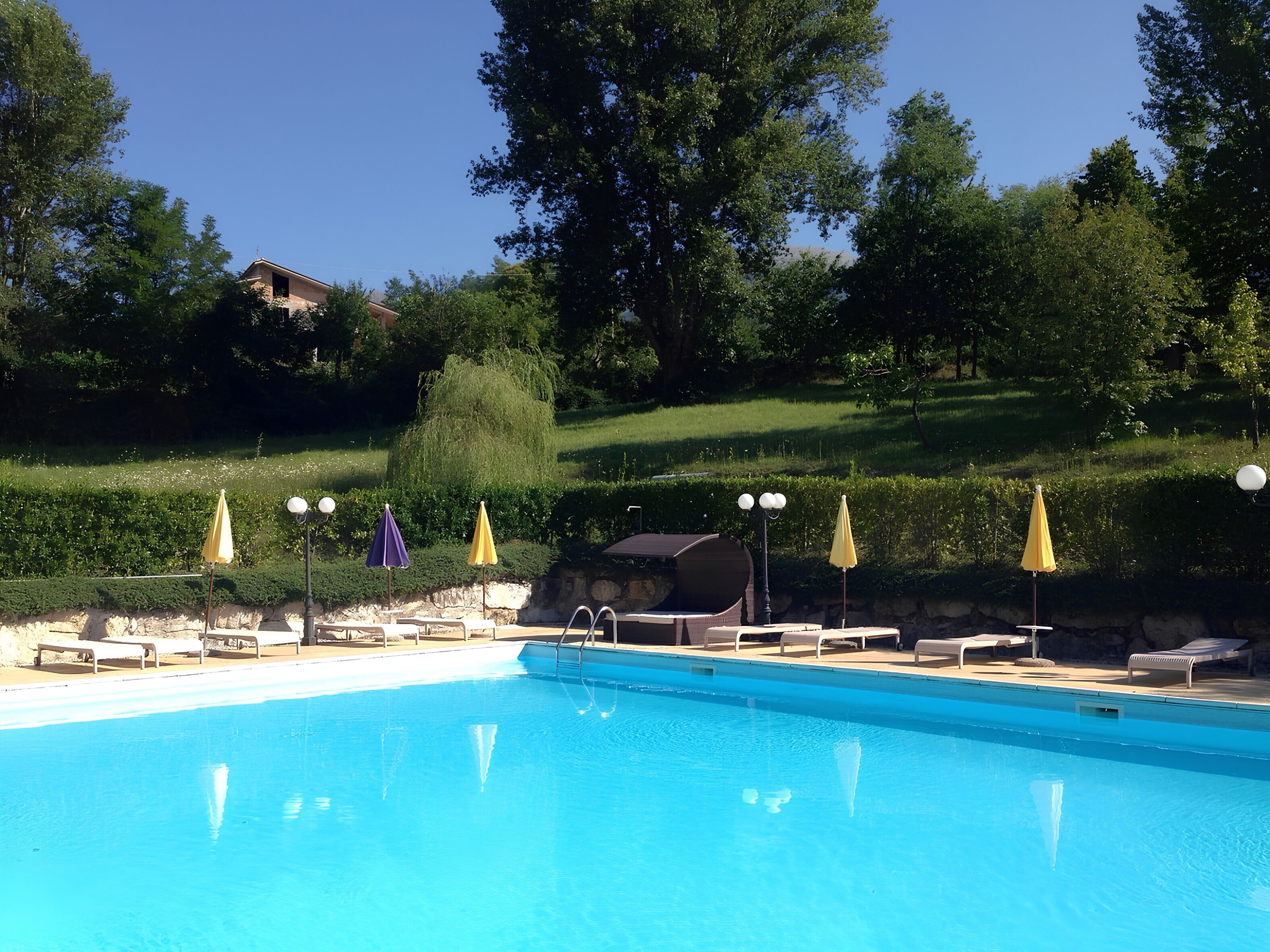 A swimming pool with lounge chairs and umbrellas, surrounded by greenery and a stone wall.