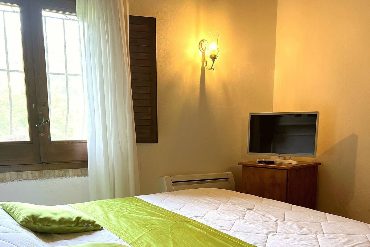 Una cama bien hecha con un corredor verde, un televisor sobre un soporte de madera y una lámpara de pared.