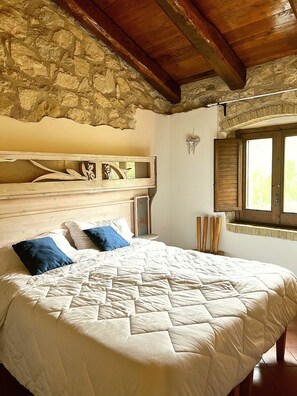 Une chambre avec un grand lit, des poutres en bois, des murs en pierre et une fenêtre.