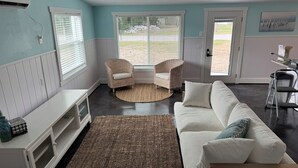 Ein Wohnzimmer mit einem weißen Sofa, zwei Korbstühlen, einem großen Fenster und einem weißen Schrank.