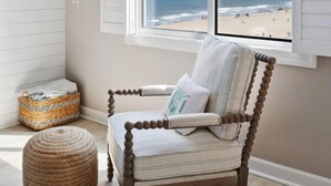 A room with a view of the beach, a wooden chair with a cushion, a woven basket, and a round ottoman.