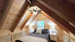 A cozy attic bedroom with a sloped ceiling, a bed with a dark-colored comforter, two bedside tables with lamps, and a window with blinds. - Robin's Nest Sleeps 6 (Mentone)