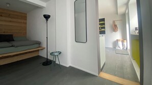 Ett modernt rum med en säng, en golvlampa, en spegel och utsikt över ett annat rum med hyllor och en väska.