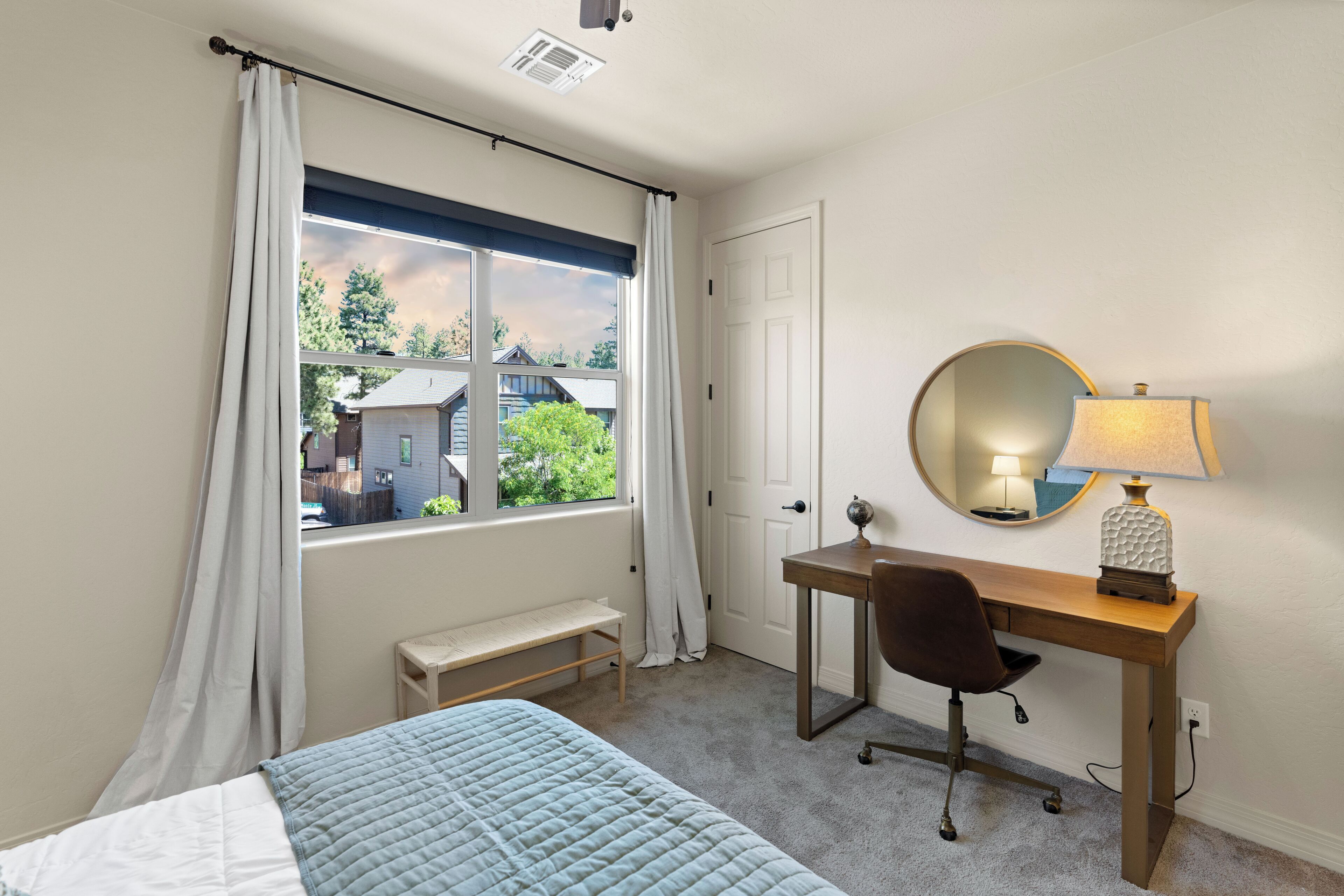 Un dormitorio con cama, escritorio, silla, lámpara, espejo y ventana con cortinas.