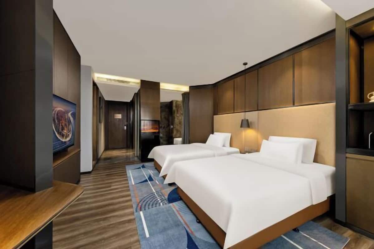 A hotel room with two beds, a wooden desk, a large mirror, and a wall-mounted TV.