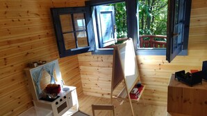 A cabin with wooden walls, a kitchen area with a stove, a table with art supplies, and a large window with a view of greenery. - Accogliente chalet sul vulcano Etna, In Sicilia. Situato a quota 1500. (Nicolosi)