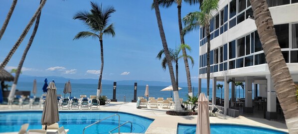 A poolside area with lounge chairs and umbrellas, overlooking a body of water and a building with large windows. - Suite una recamara en la playa (Bucerías)
