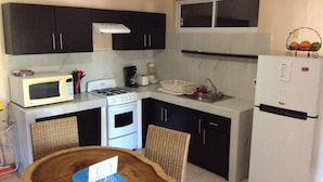 Cocina compacta con microondas, estufa, fregadero y refrigerador. Incluye una silla de mimbre y una pequeña mesa de comedor con un mantel individual.