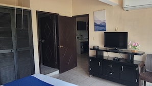Un dormitorio con cama, televisor sobre un soporte negro, una silla y un armario.