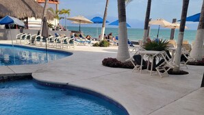 A pool area with lounge chairs and umbrellas, a clear view of the ocean, and a thatched-roof structure in the background. - Estudio frente a la playa (Bucerías)