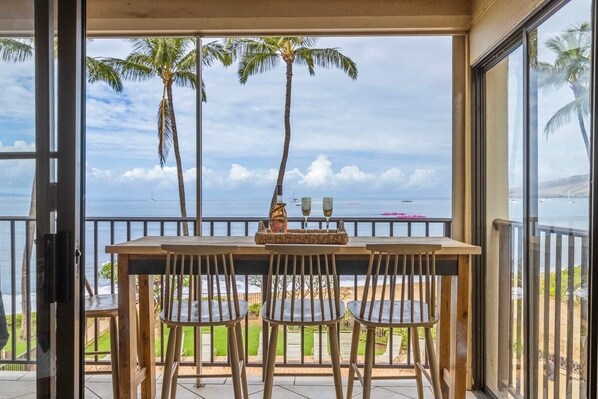 A balcony with a table and chairs, overlooking a beach with palm trees and the ocean.
