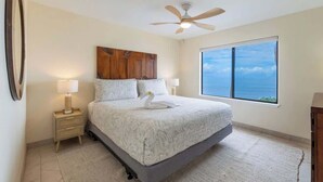 Ein Schlafzimmer mit einem Bett, einem Deckenventilator, einem Nachttisch, einer Lampe, einem Fenster mit Blick und einer Wanduhr.