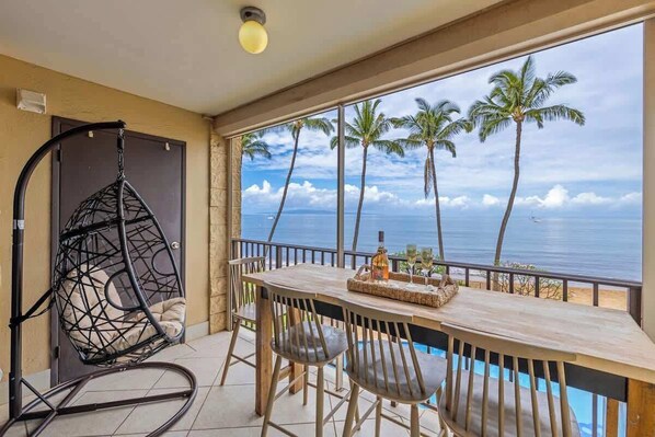Un balcón con vista al océano y palmeras, una silla colgante y una mesa con una bandeja.
