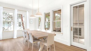 Une salle à manger lumineuse, avec une table en bois et des chaises blanches, de grandes fenêtres et une cheminée.