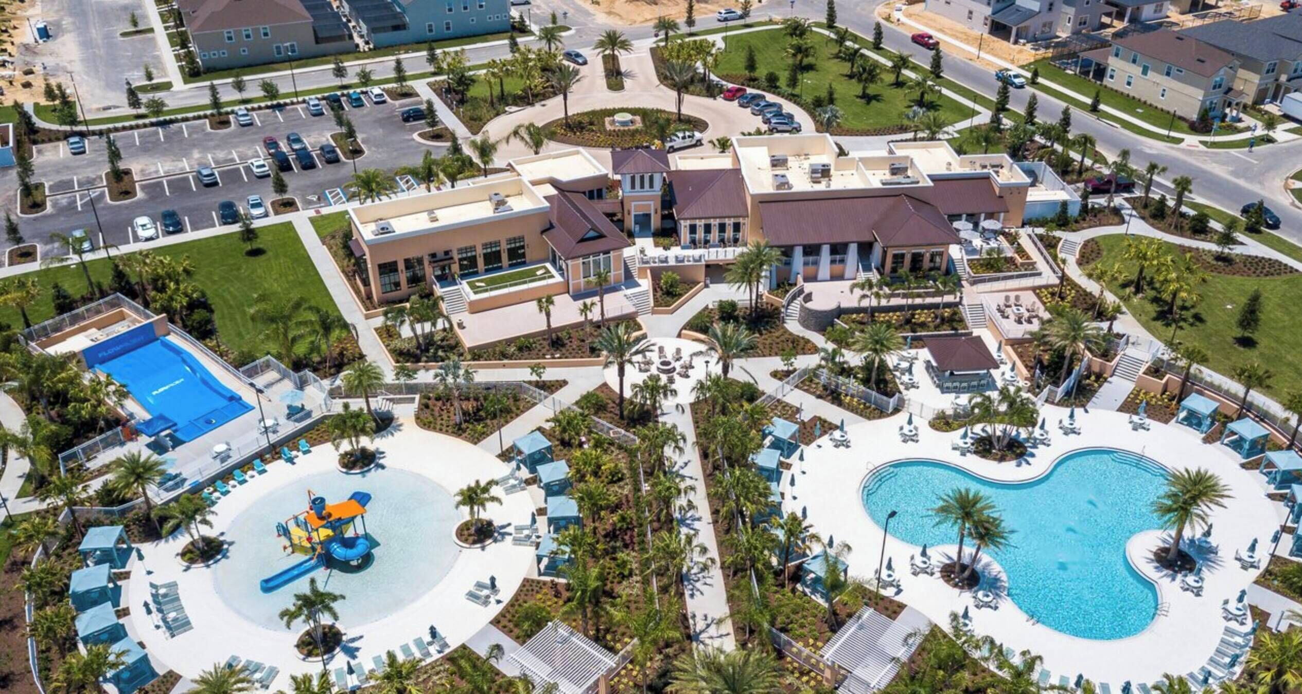 Vista aérea de un complejo turístico con una amplia piscina central, varias edificaciones y un estacionamiento.