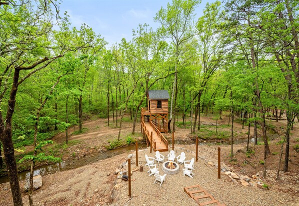A forest cabin with a deck and a fire pit surrounded by chairs.