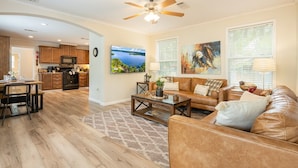 A spacious living room with a leather sofa, a coffee table, and a TV. There is a kitchen in the background with wooden cabinets and a microwave. - Eagle`s Cove · Waterfront Amazing View, King Bed, Deck (Hemphill)