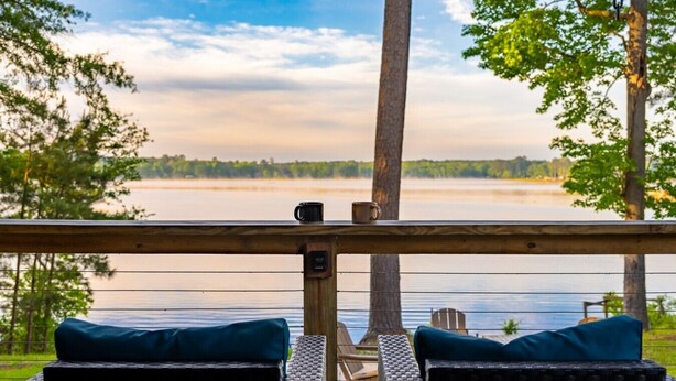 A wooden deck with two wicker chairs and cushions, overlooking a calm lake with trees in the background. - Eagle`s Cove · Waterfront Amazing View, King Bed, Deck (Hemphill)