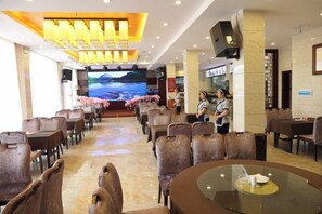 A hotel dining area with multiple tables and chairs, a large screen displaying a scenic view, and a staff member standing near the entrance. - Xinning Xiaobaihua Resort Hotel (Xinning)