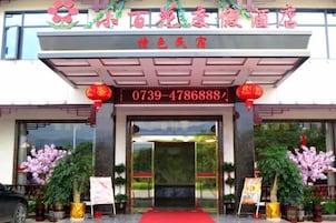 Entrada de hotel con alfombra roja, puertas de cristal grandes y plantas en macetas.