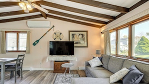 Ein Wohnzimmer mit Holzbalkendecke, einem Fernseher, einer Couch und einem Esstisch mit Stühlen.