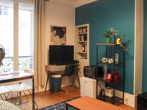Un espacio habitable compacto con escritorio, televisor, estantería y refrigerador pequeño.