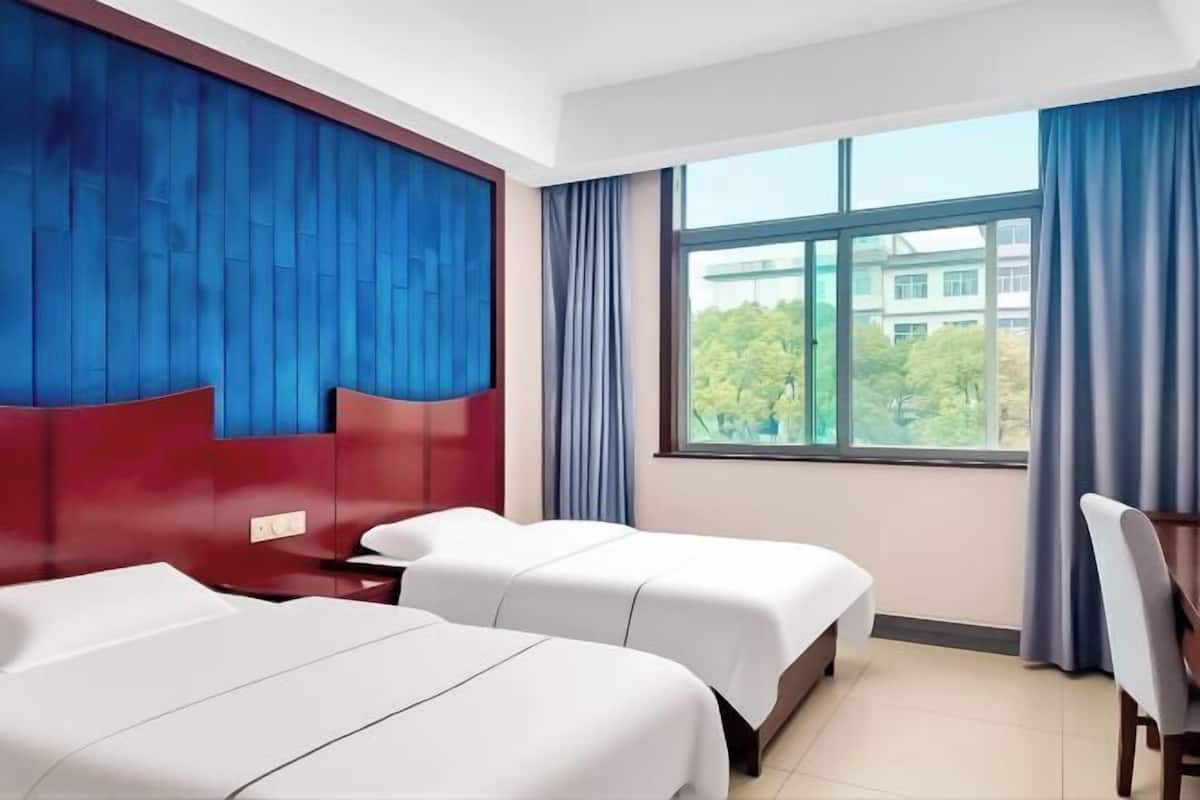 Een hotelkamer met twee bedden, een bureau en een groot raam met uitzicht op gebouwen en bomen.